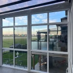 Inside a glass enclosure with a clear roof and white-framed windows overlooking open fields